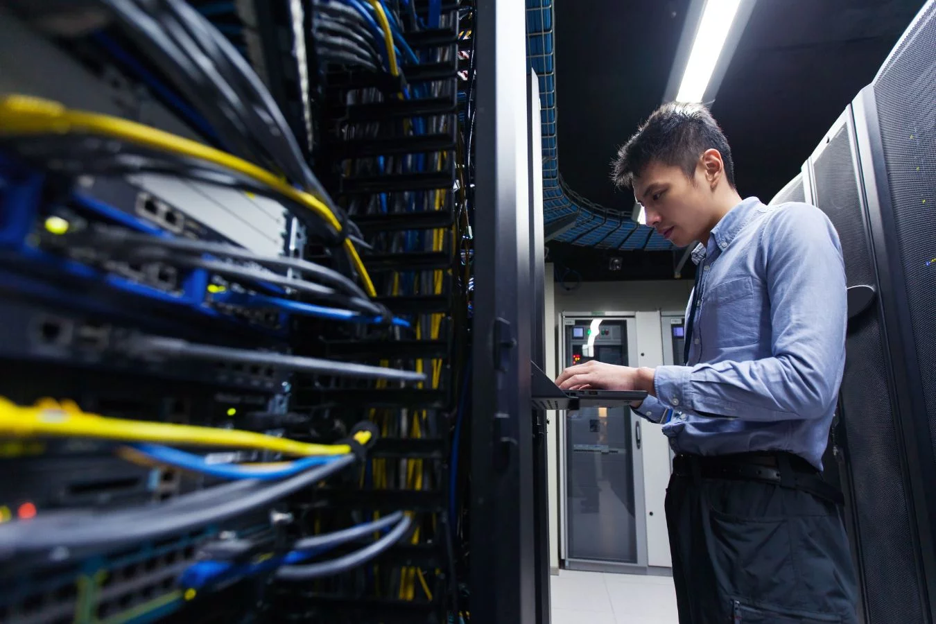 In a server room brimming with network cables and equipment, a person in a blue shirt diligently works on a laptop, embodying the essence of Who We Are at RamcoTek Consulting services.