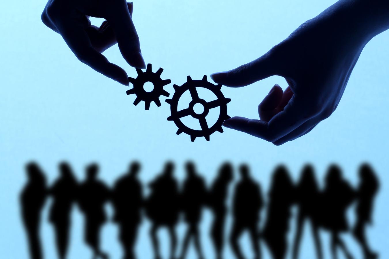 Silhouetted hands connecting gears with a blurred group of people in the background, symbolizing teamwork and collaboration in Managed IT Services.