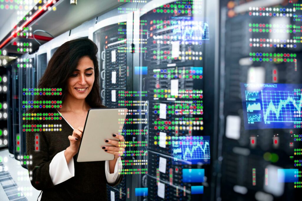 A professional woman using a tablet while standing in a data center with server racks and performance graphs in the background, providing seamless system integration services.