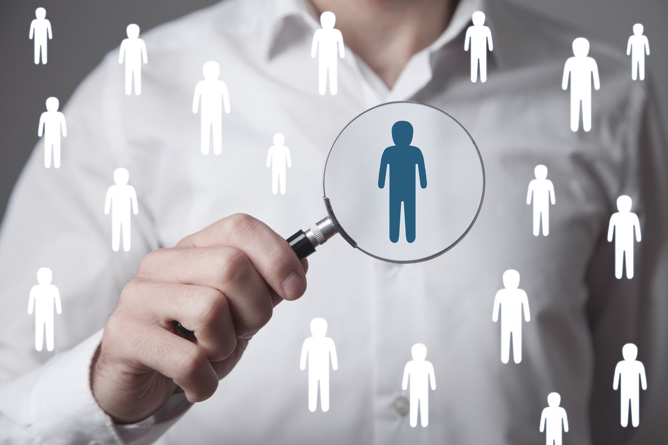 Man holding a magnifying glass over a selected figure among many identical silhouettes for recruitment purposes.