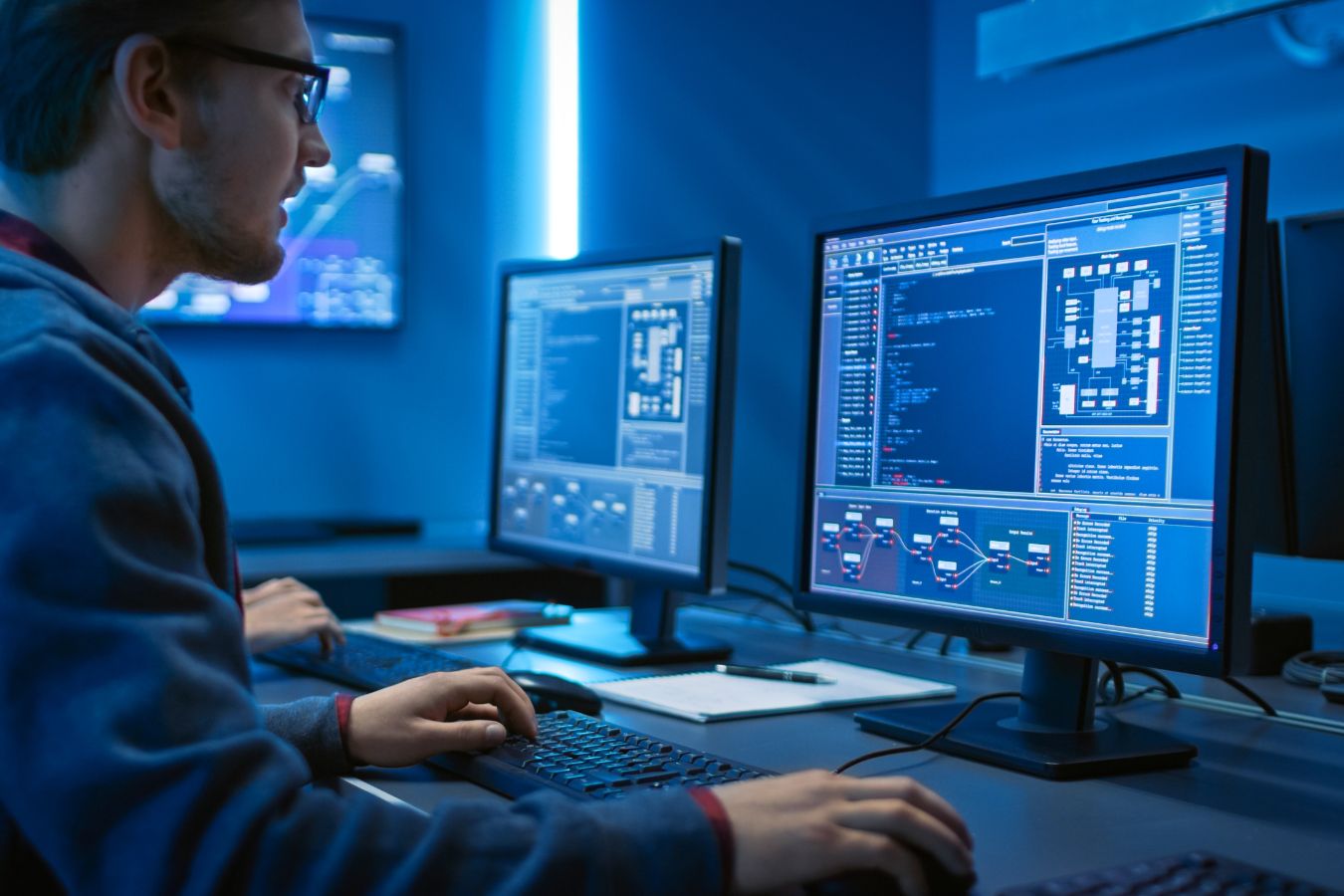 A software developer working on code at a dual-monitor workstation in a dimly lit room.