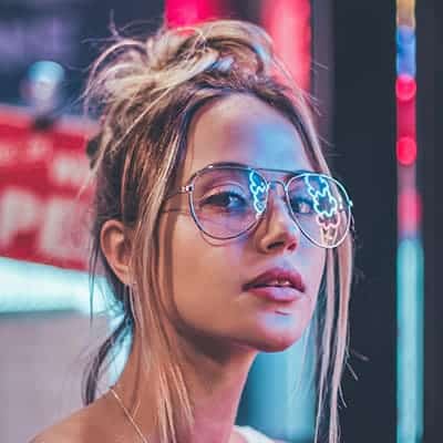 Young woman wearing reflective sunglasses with neon lights visible in the reflection.